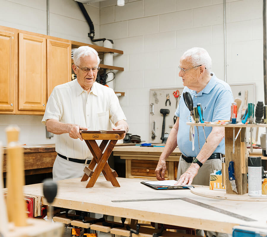 Two senior men crafting in a woodworking shop with tools and wooden furniture projects.