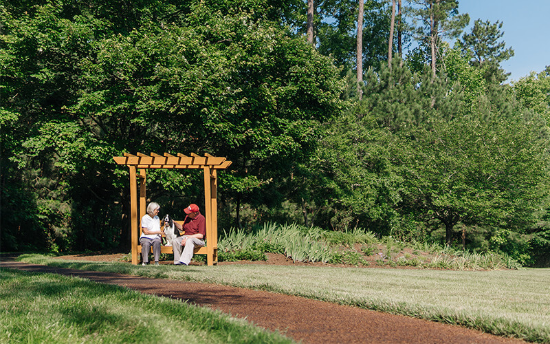 Two seniors sitting under a wooden pergola in a lush garden area.