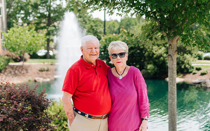 Peaceful Afternoon by the Pond Elderly couple smiling by a pond with a fountain in a serene landscaped garden.