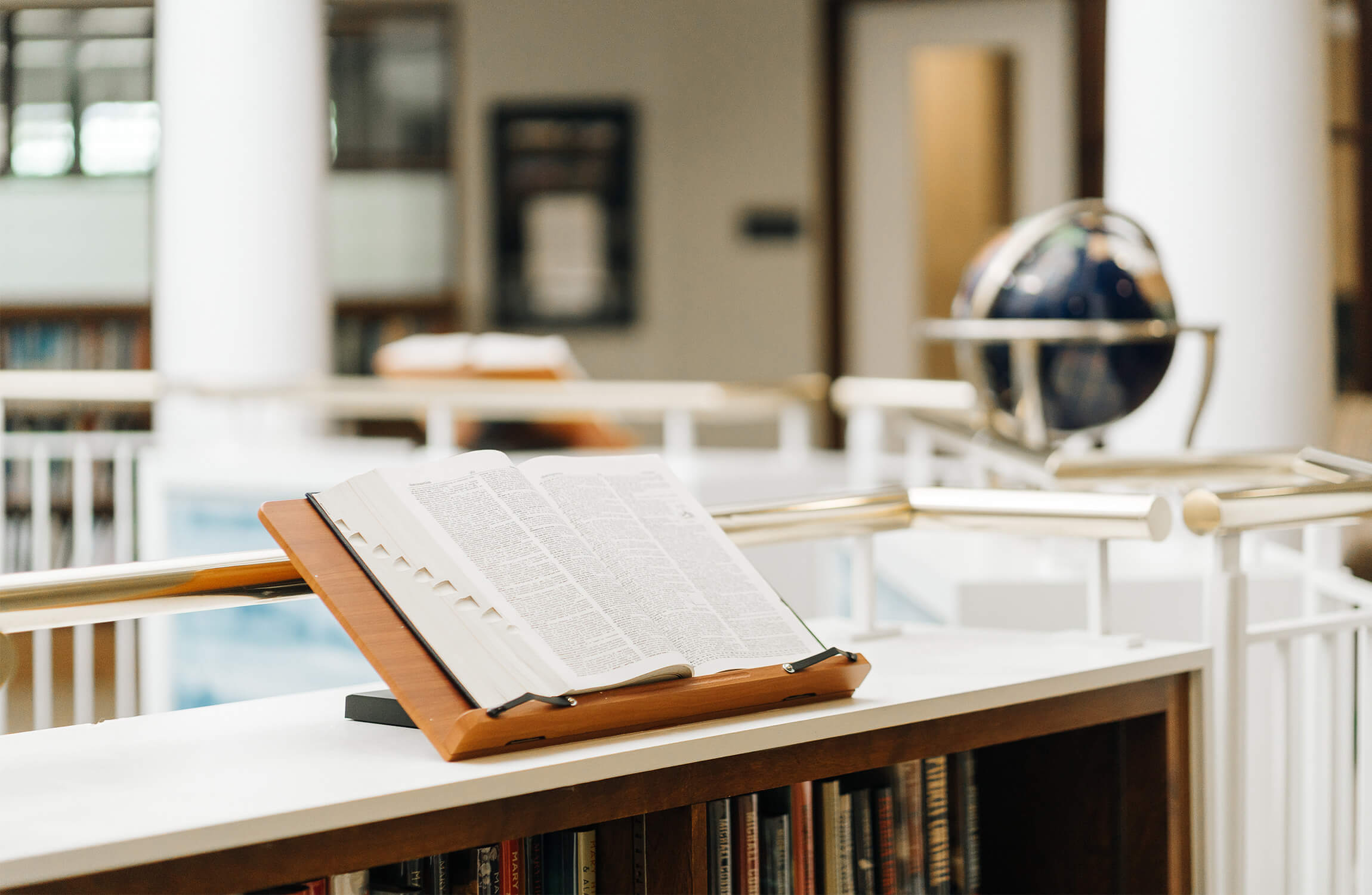 Open book on a wooden stand in a well-lit library with a globe in the background.