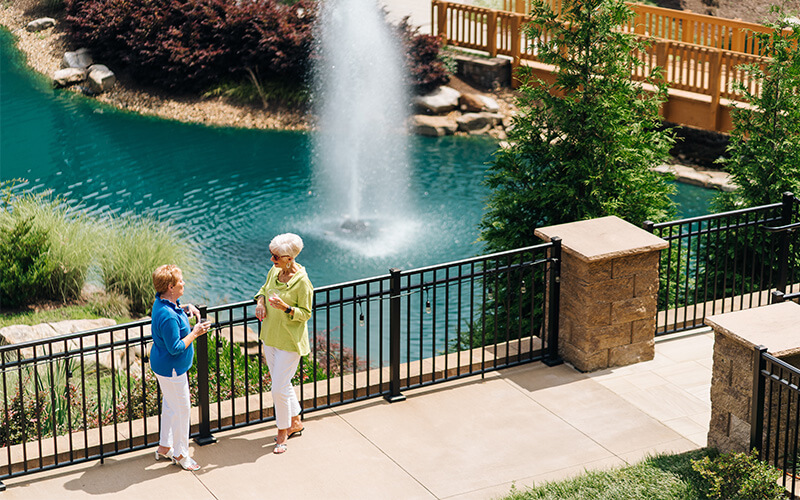 Two women talking by a pond with a fountain in a senior living community.