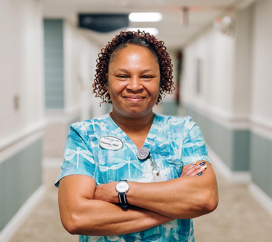 Smiling nurse in scrubs with folded arms in a senior living community hallway.