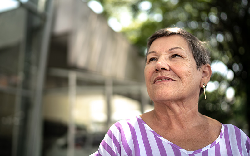 Smiling elderly woman outside a building with glass windows and trees in the background.