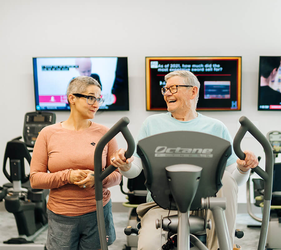 Elderly man on exercise machine with supportive woman by his side in a fitness room.