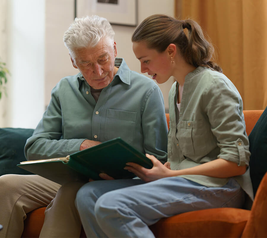 Elderly man and young woman sitting on a sofa, looking at a photo album together.