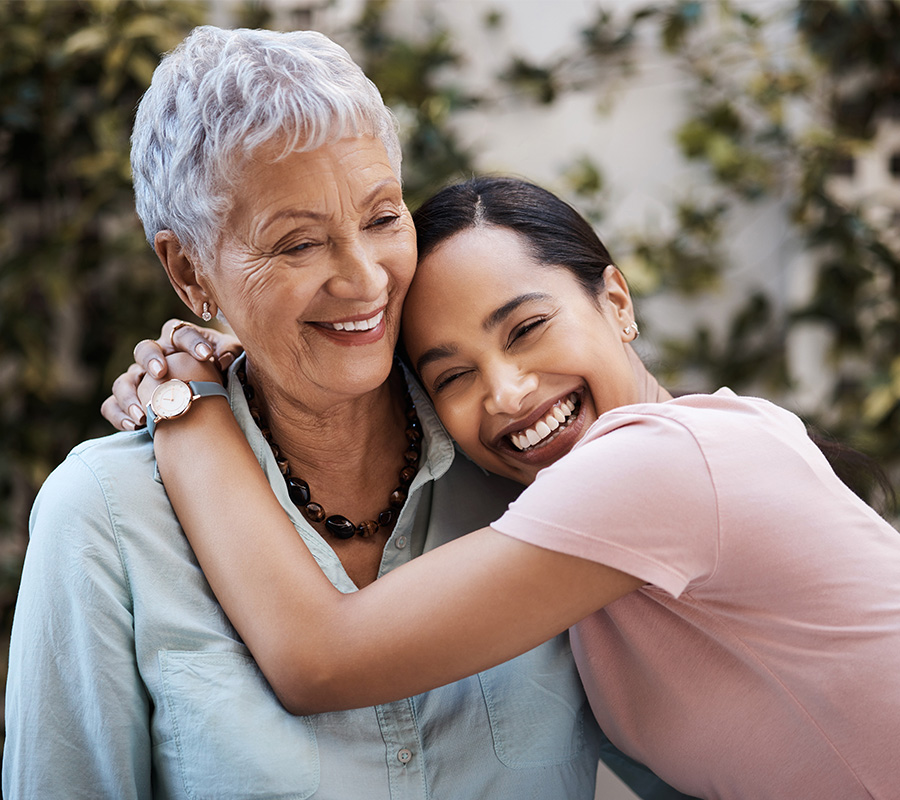 Elderly woman hugged by young woman, both smiling warmly in garden setting.