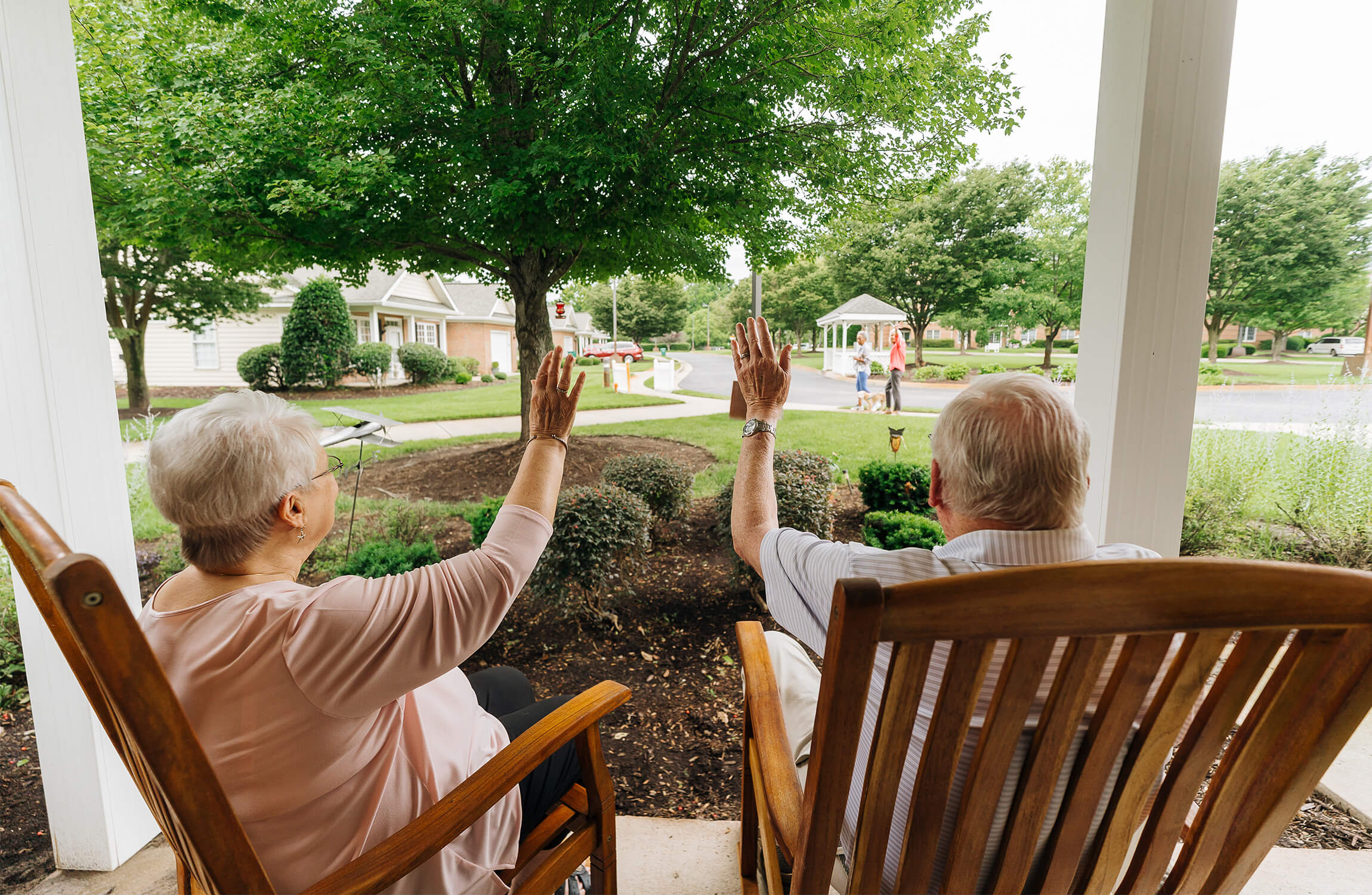 Seniors sitting on porch waving at people walking in a community with units and greenery.