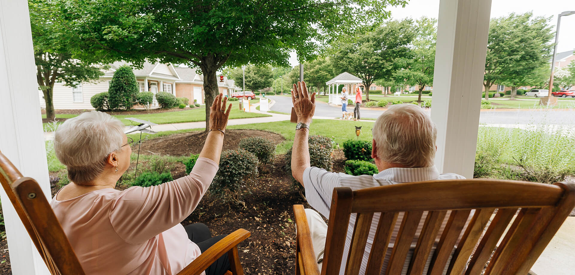 Older couple waving from porch at people walking in community garden.