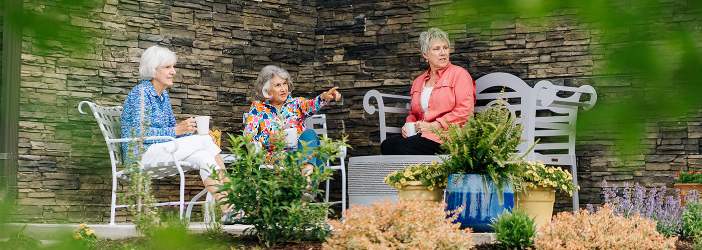 Three women enjoying coffee on a patio with lush greenery and stone walls.