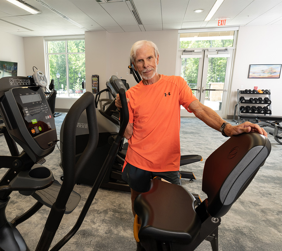 Senior man in orange shirt using exercise equipment in bright fitness center.