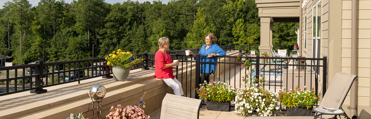 Two people enjoy coffee on a sunlit balcony at a senior living community.