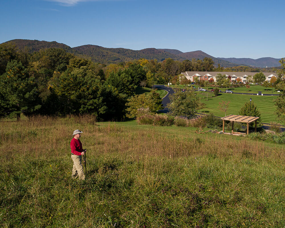 Person hiking in a field with senior living units and mountains in the background.