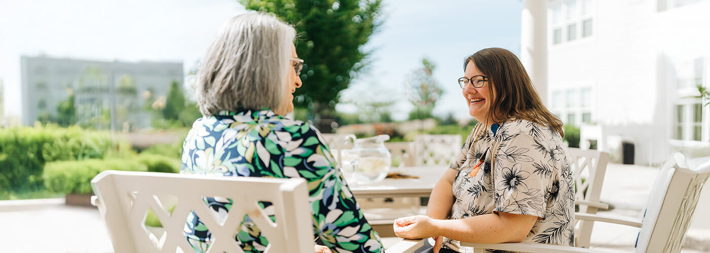 Two women talking at a table outdoors in a senior living community.