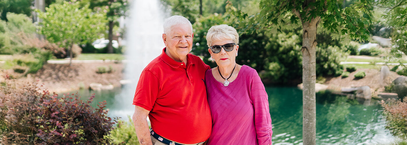 Smiling elderly couple standing by a pond in a scenic garden setting.