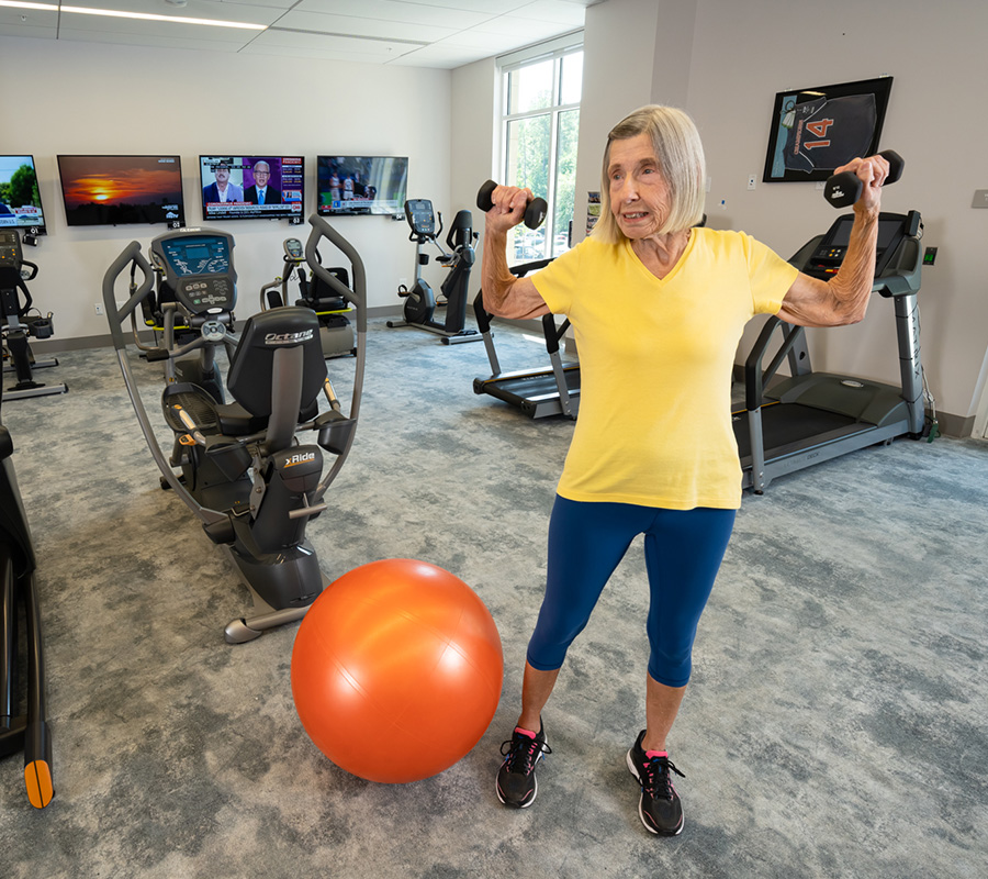 Elderly woman exercising with weights in a fitness room with gym equipment and TVs.