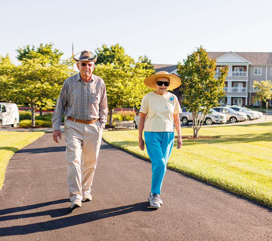 Two seniors walking on a sunny path near a residential building.