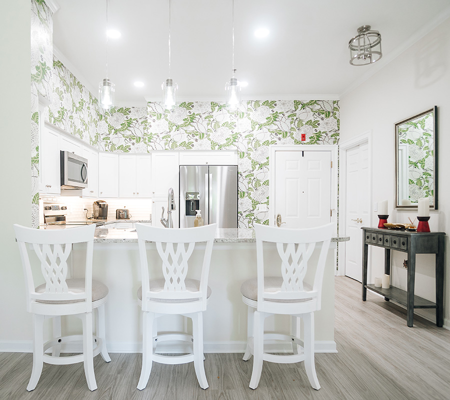Bright kitchen with floral wallpaper, white cabinets, and bar stools in a senior living unit.