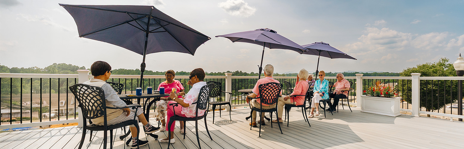 Residents relaxing on a sunny patio under umbrellas at a senior living community.