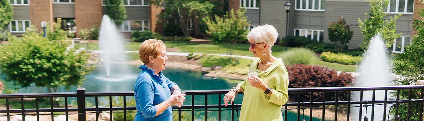 Two senior women stand outside conversing against a fence overlooking a pond with a fountain, and senior community apartments.