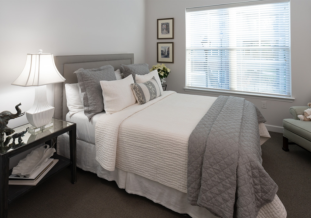 Cozy bedroom with a plush bed, elegant lamp, and bright window in a senior living unit.