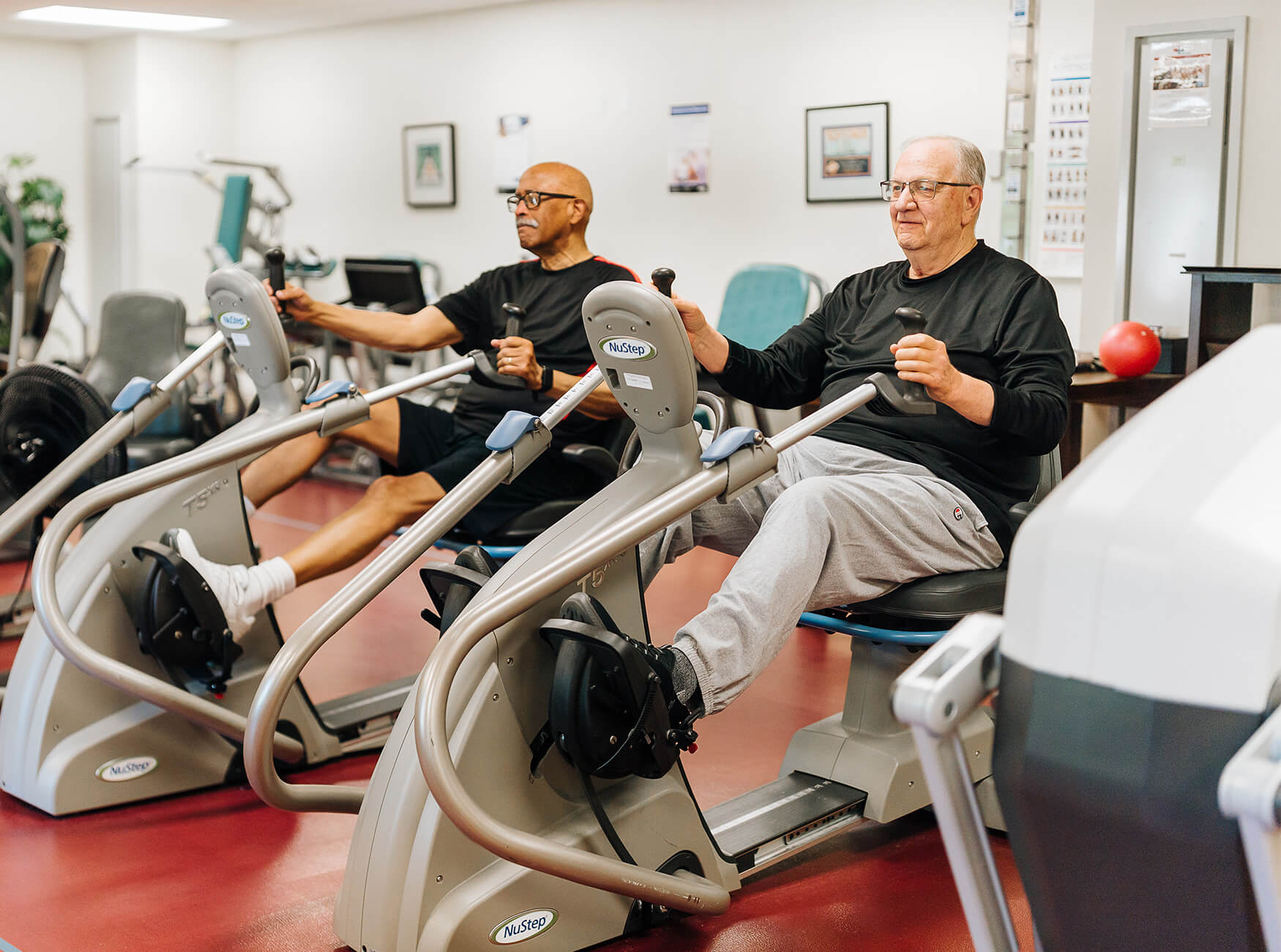Two seniors exercising on stationary cycling machines in a fitness room.