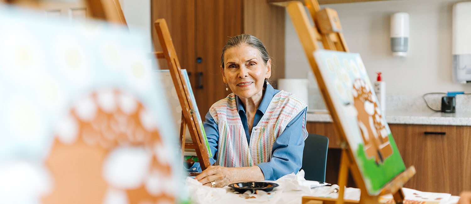 Elderly woman painting at easel in a communal art room.