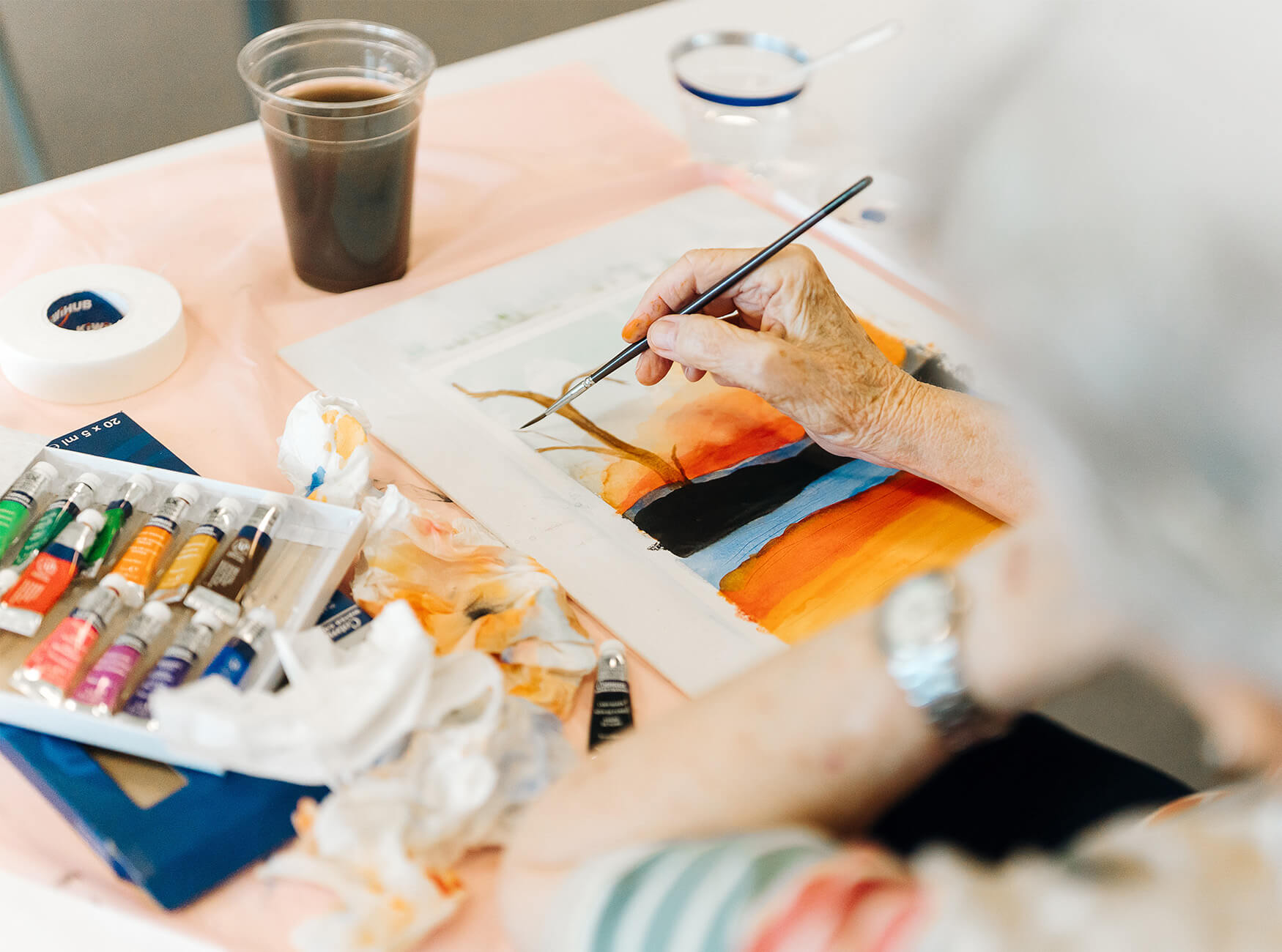 Senior person painting a vibrant landscape at a workshop table.