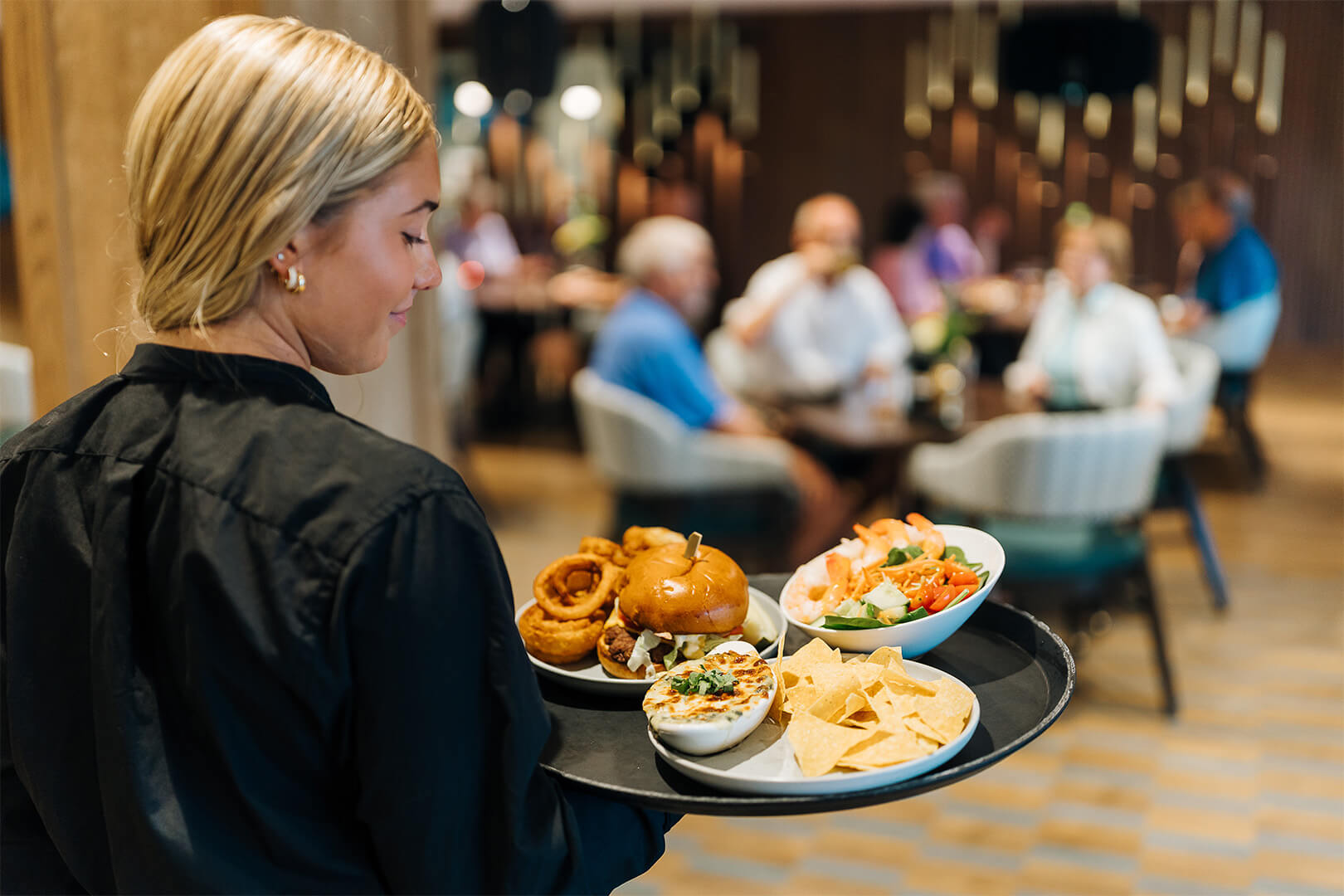 Server carrying a tray with diverse meals in a communal dining area.