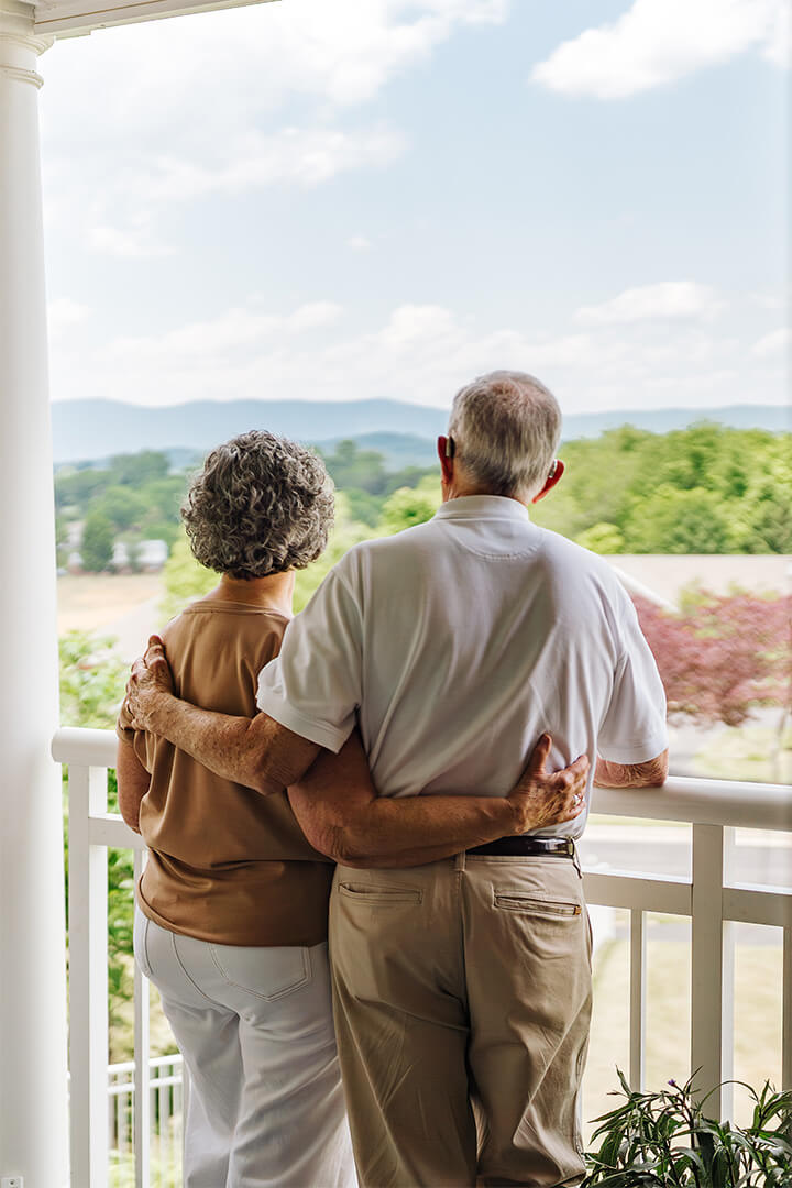 Elderly couple embracing on a porch, enjoying a scenic view of mountains and greenery.