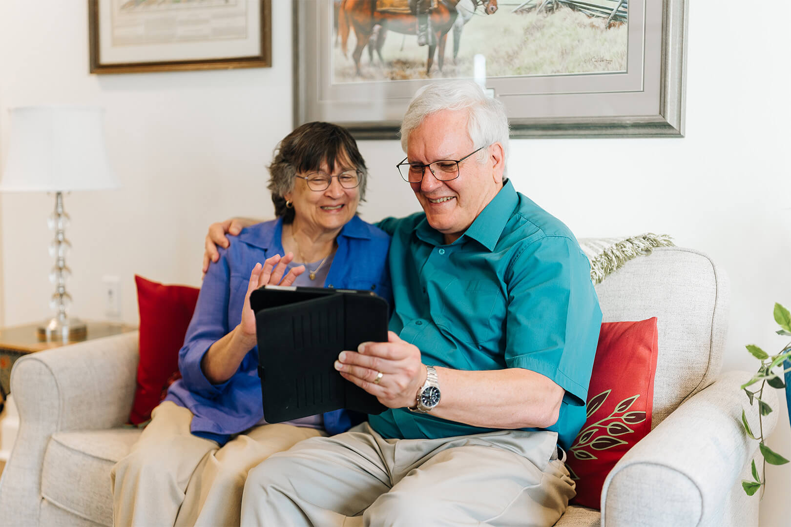 Smiling elderly couple sitting on a couch looking at a tablet in a cozy living room.