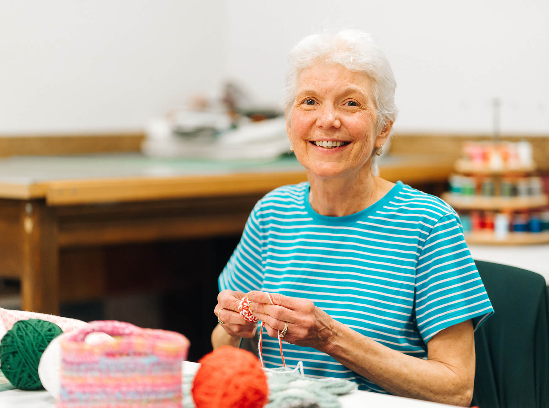 Elderly woman smiling while knitting at a table with colorful yarn.