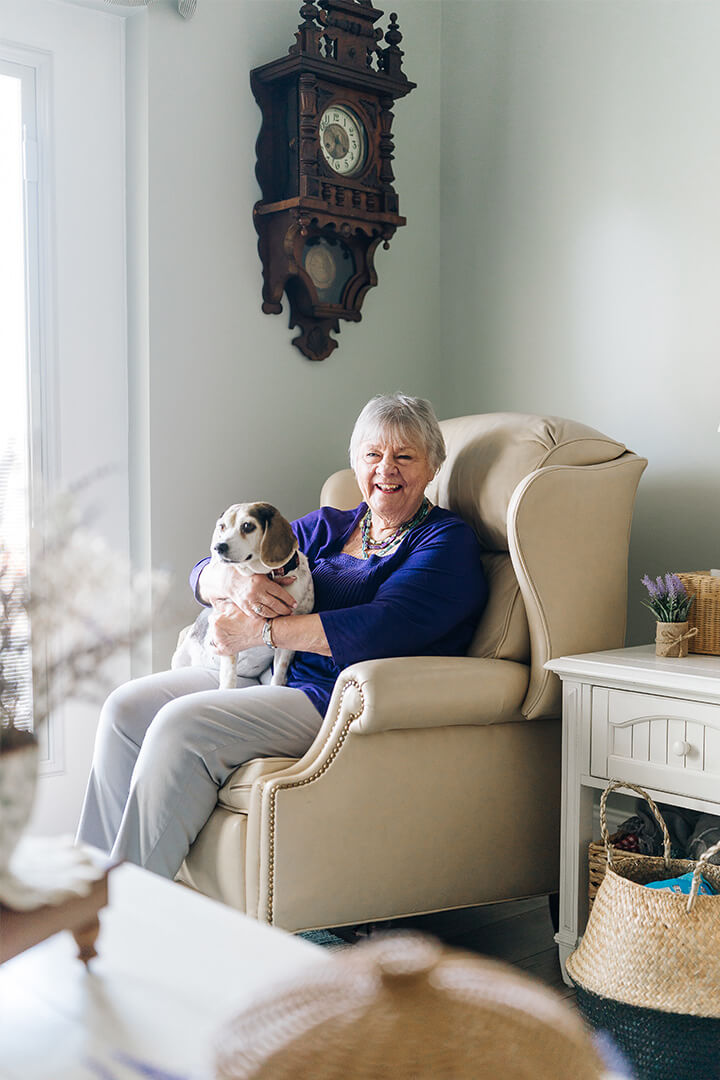 Elderly woman in a cozy chair holds a small dog, smiling warmly in a bright room.
