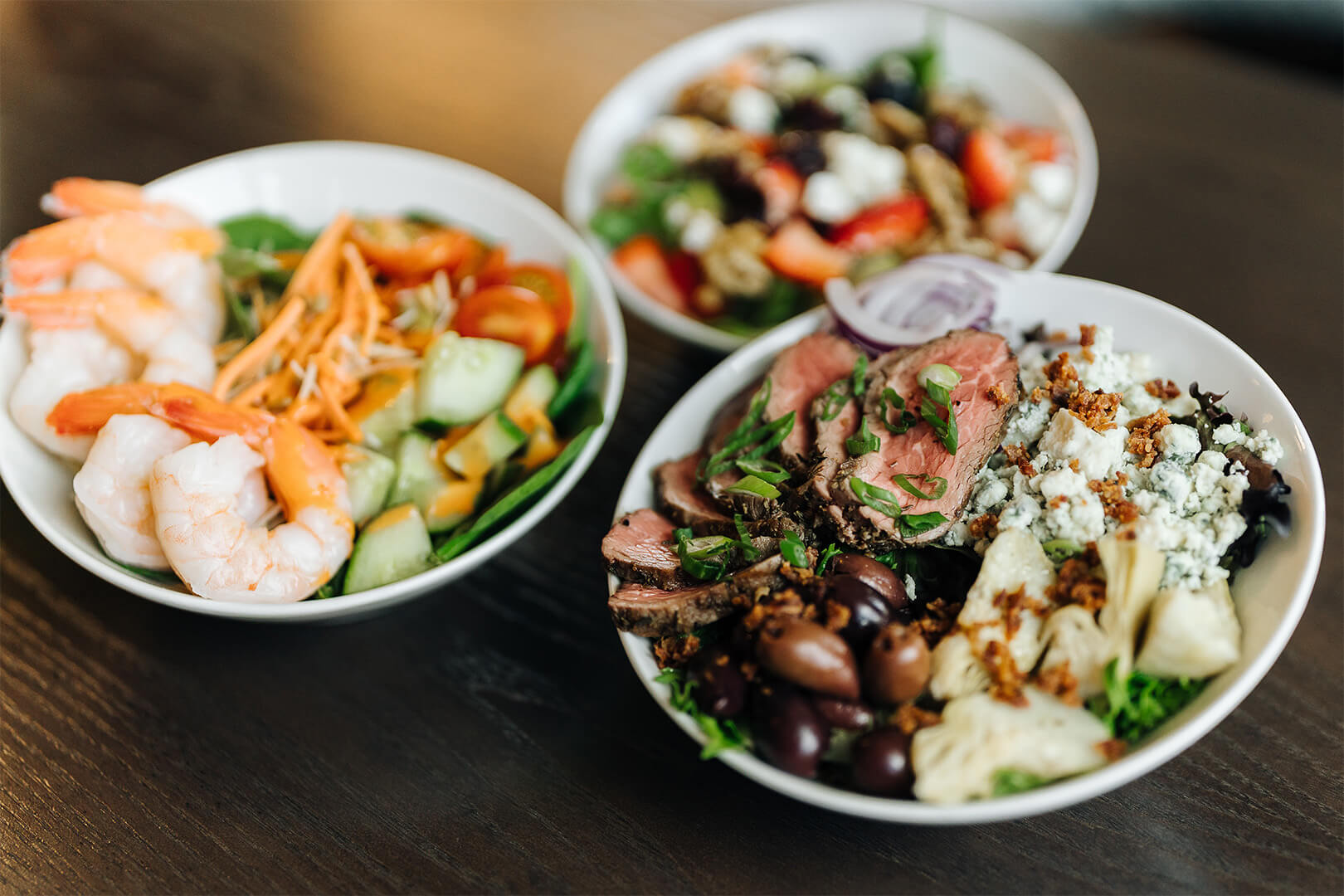 Three bowls of fresh salads with shrimp, greens, and assorted toppings on a wooden table.