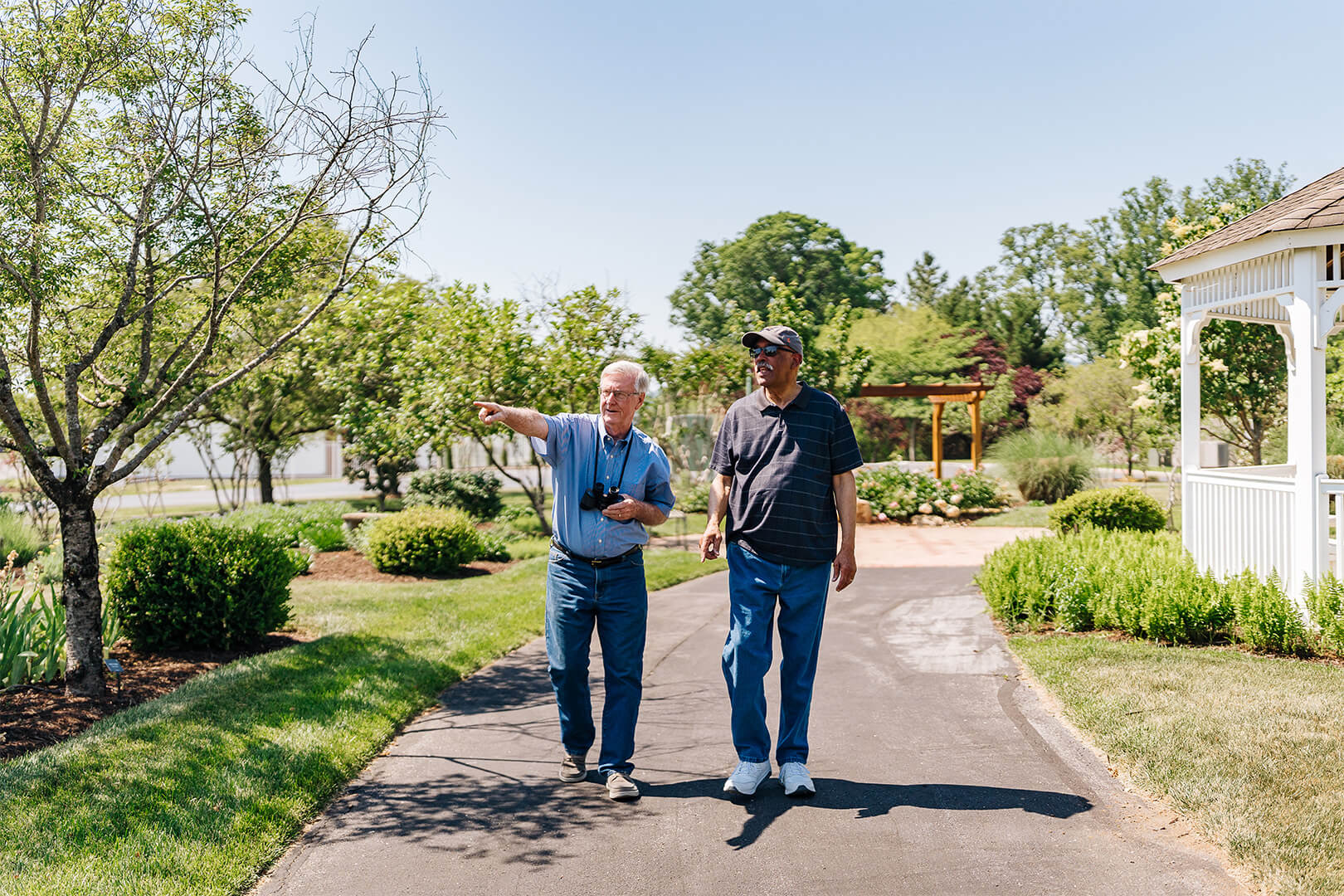 Two men walking on a path through a garden, one pointing, in a senior living community.