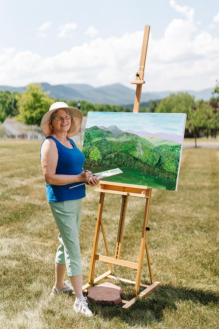 Smiling woman painting outdoors on a sunny day with easel in the background.
