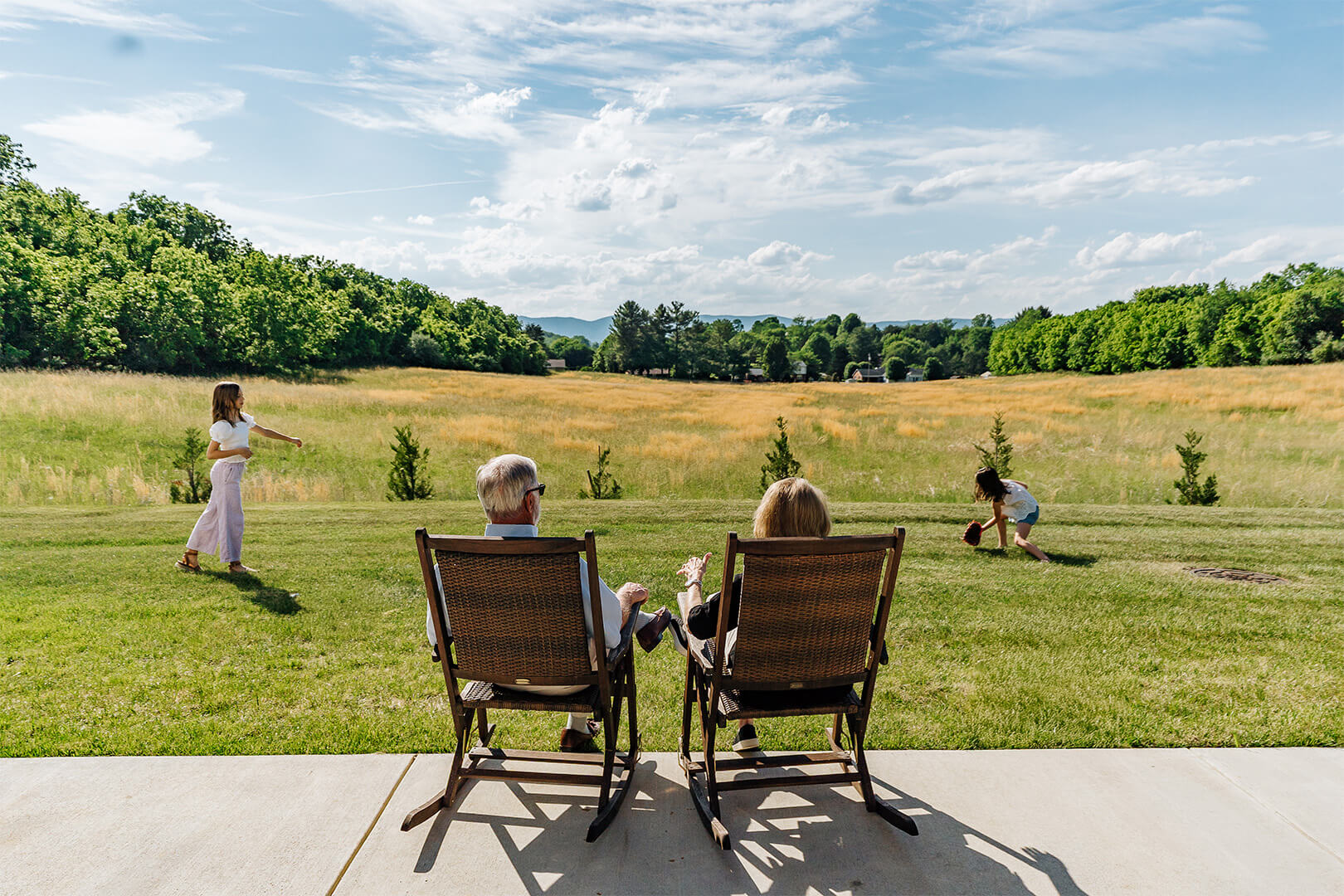 Two seniors sit in rocking chairs overlooking a field while children play on a sunny day.