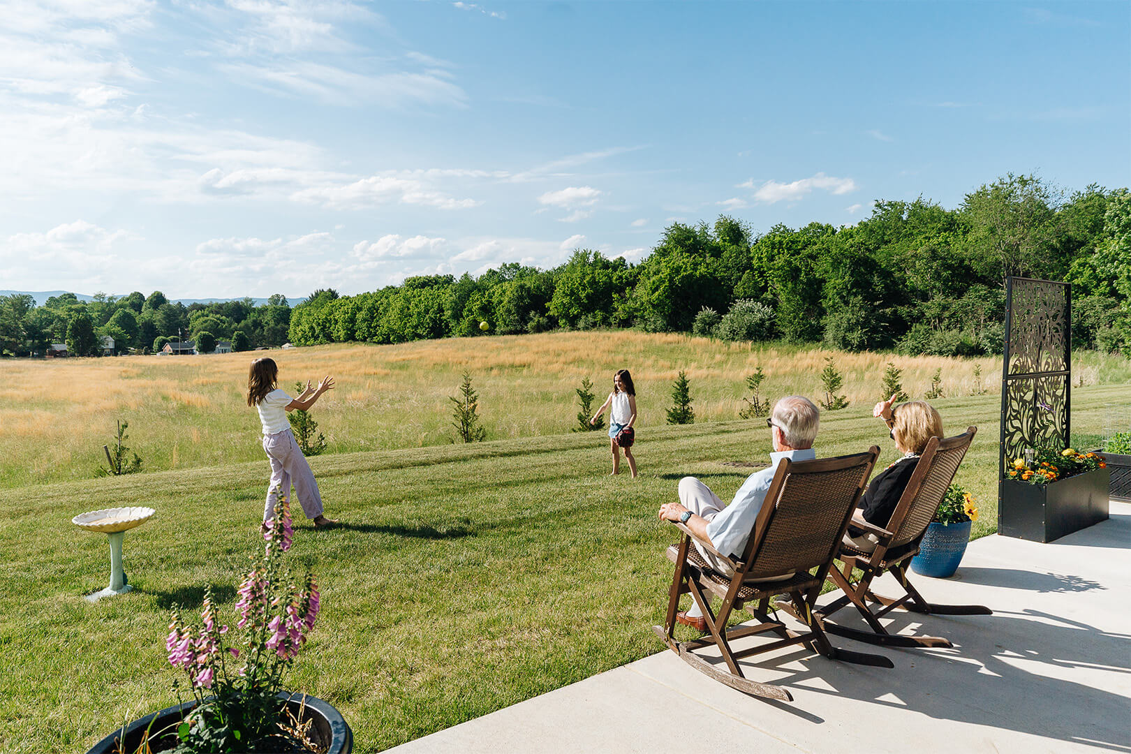 Grandparents watching children play catch on a sunny day in an open grassy field.