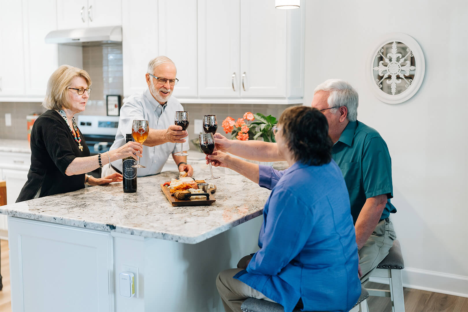 Four senior residents enjoying wine and snacks in a kitchen setting.