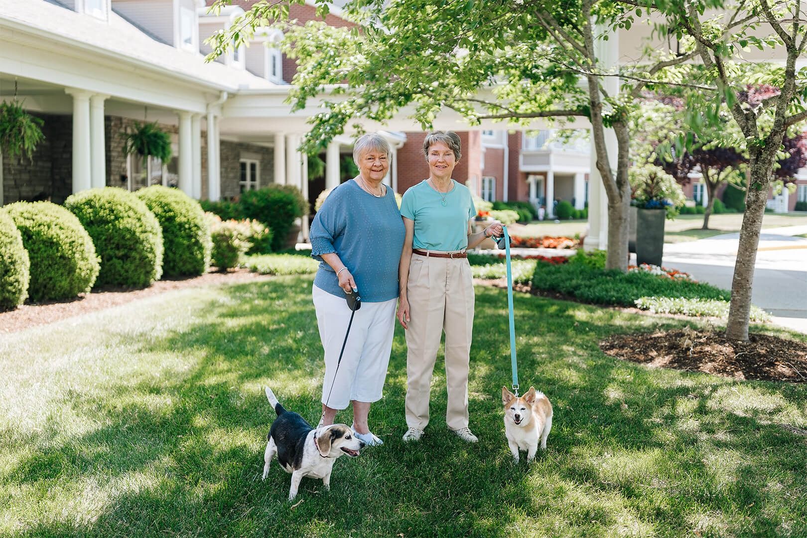 Two smiling women with dogs in front of a large building and trees.