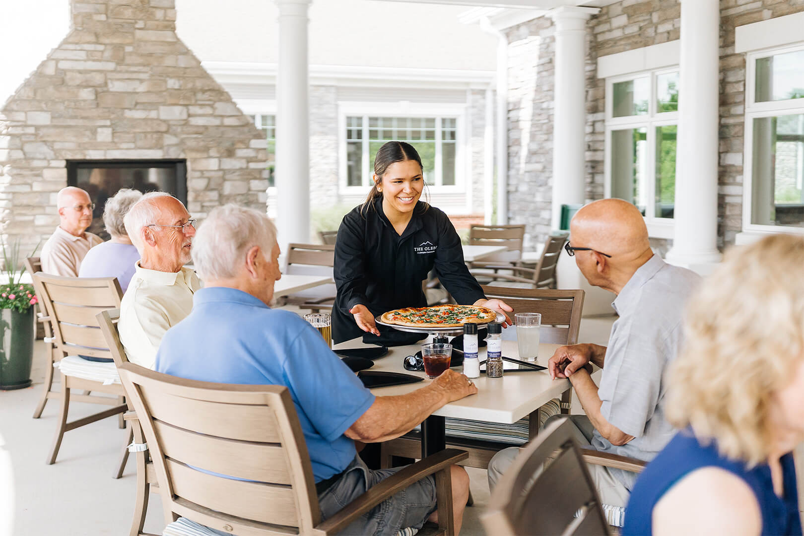 Waitress serving pizza to smiling seniors at a dining area in a senior living community.