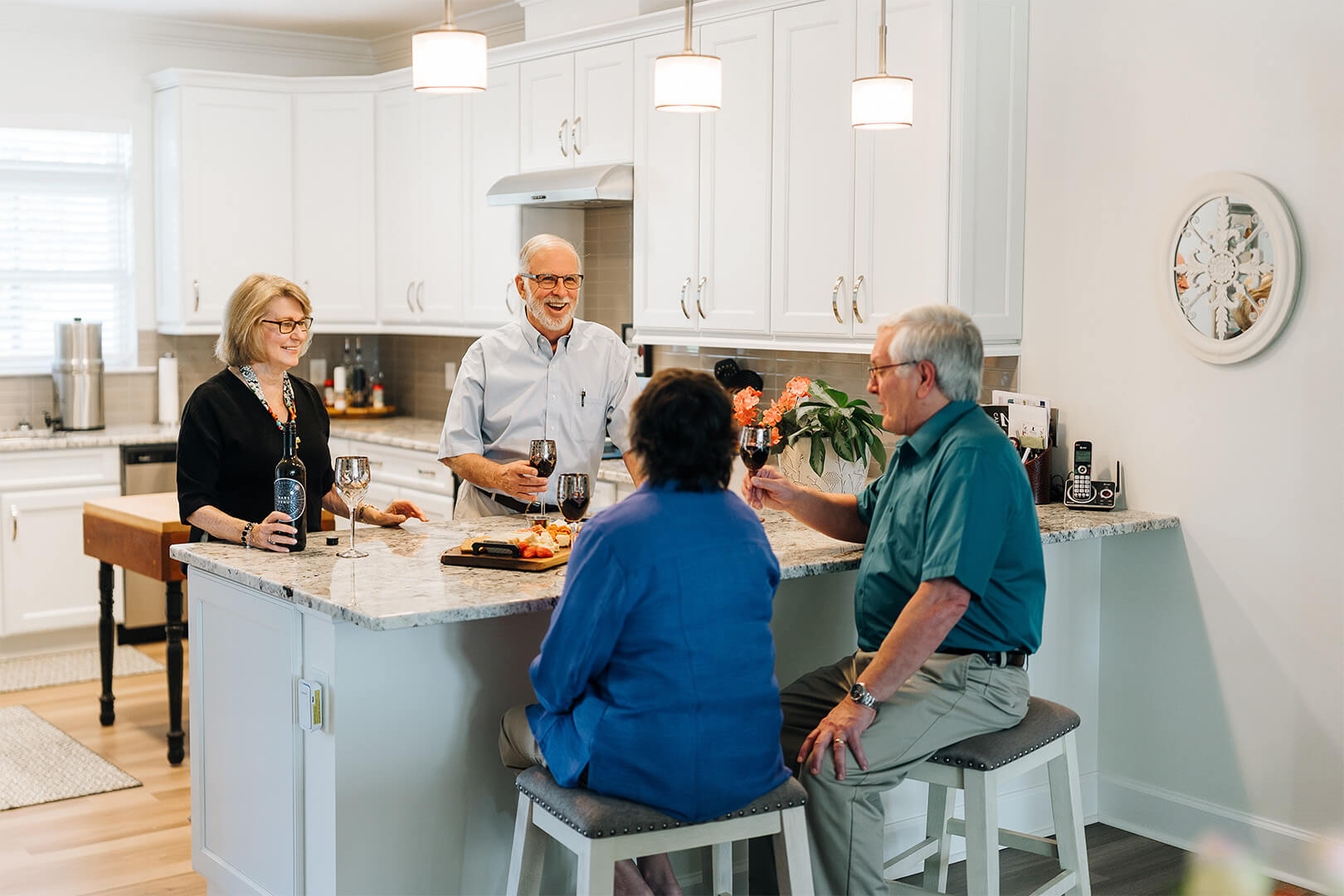 Four seniors enjoying wine and snacks in a bright kitchen with white cabinets.
