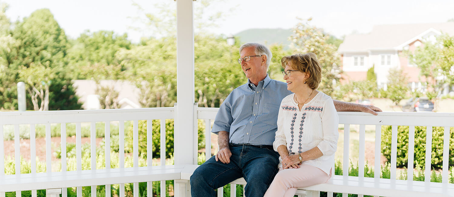 Senior couple sitting on gazebo bench in a garden at a community with housing units in view.