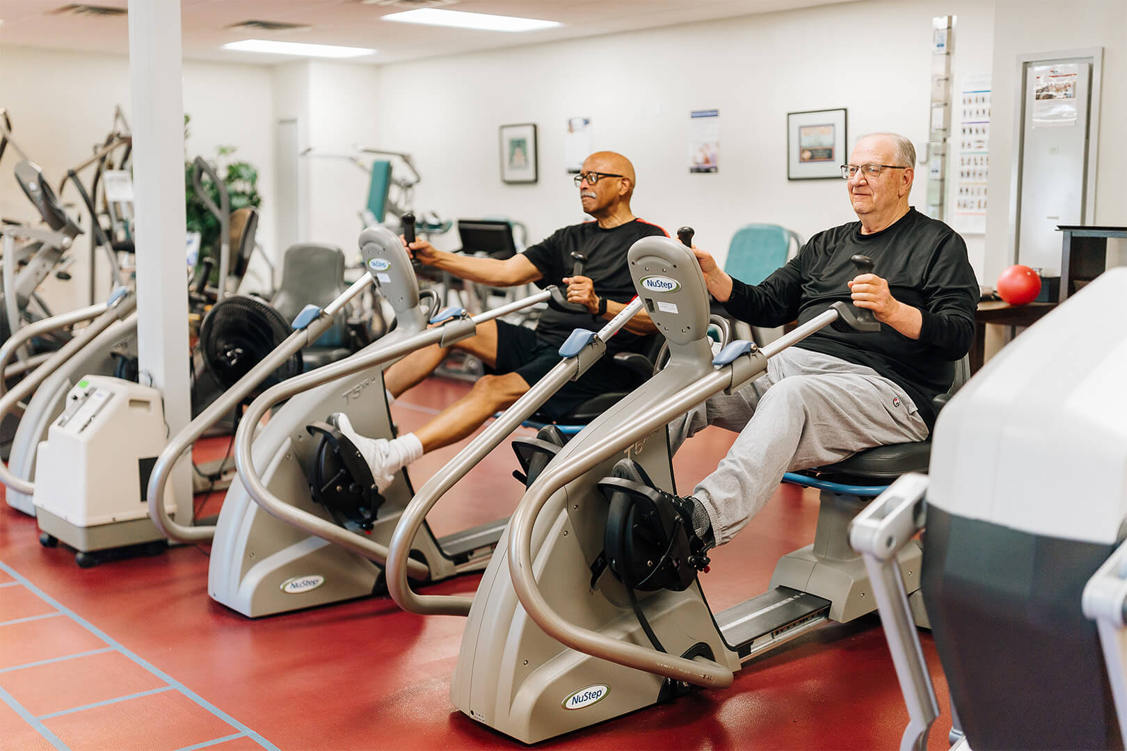 Two elderly men exercising on recumbent cross trainers in a fitness room.