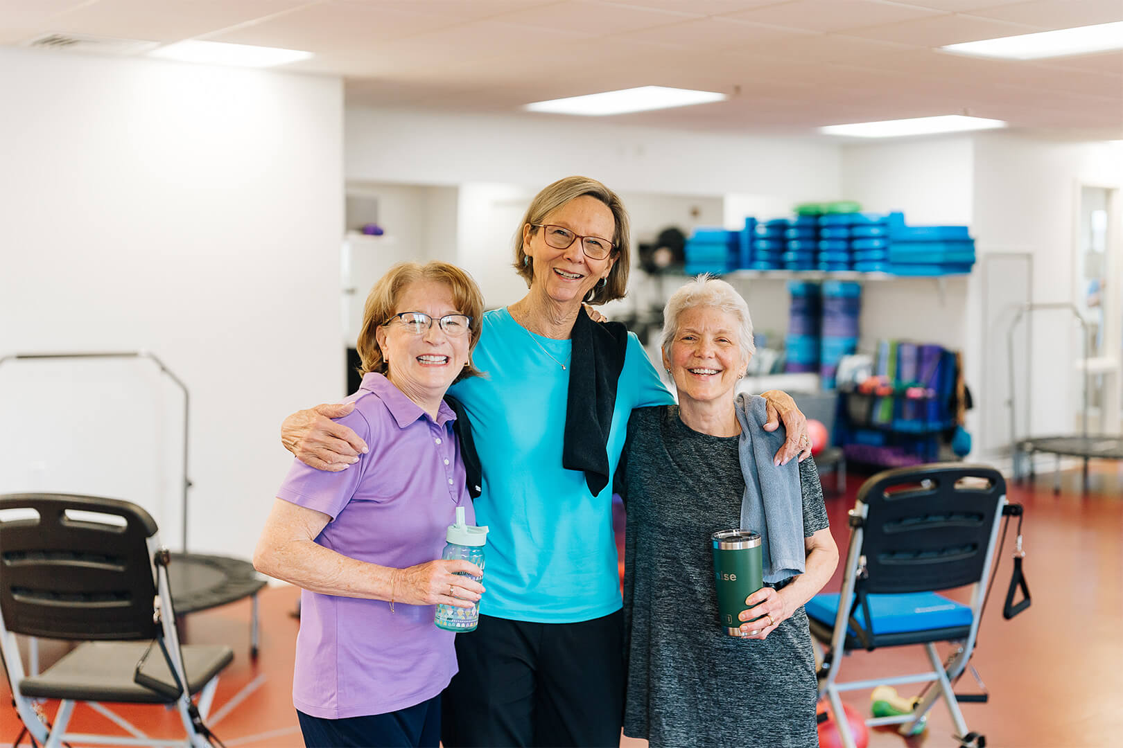 Three smiling senior women in a fitness room, holding water bottles and embracing.