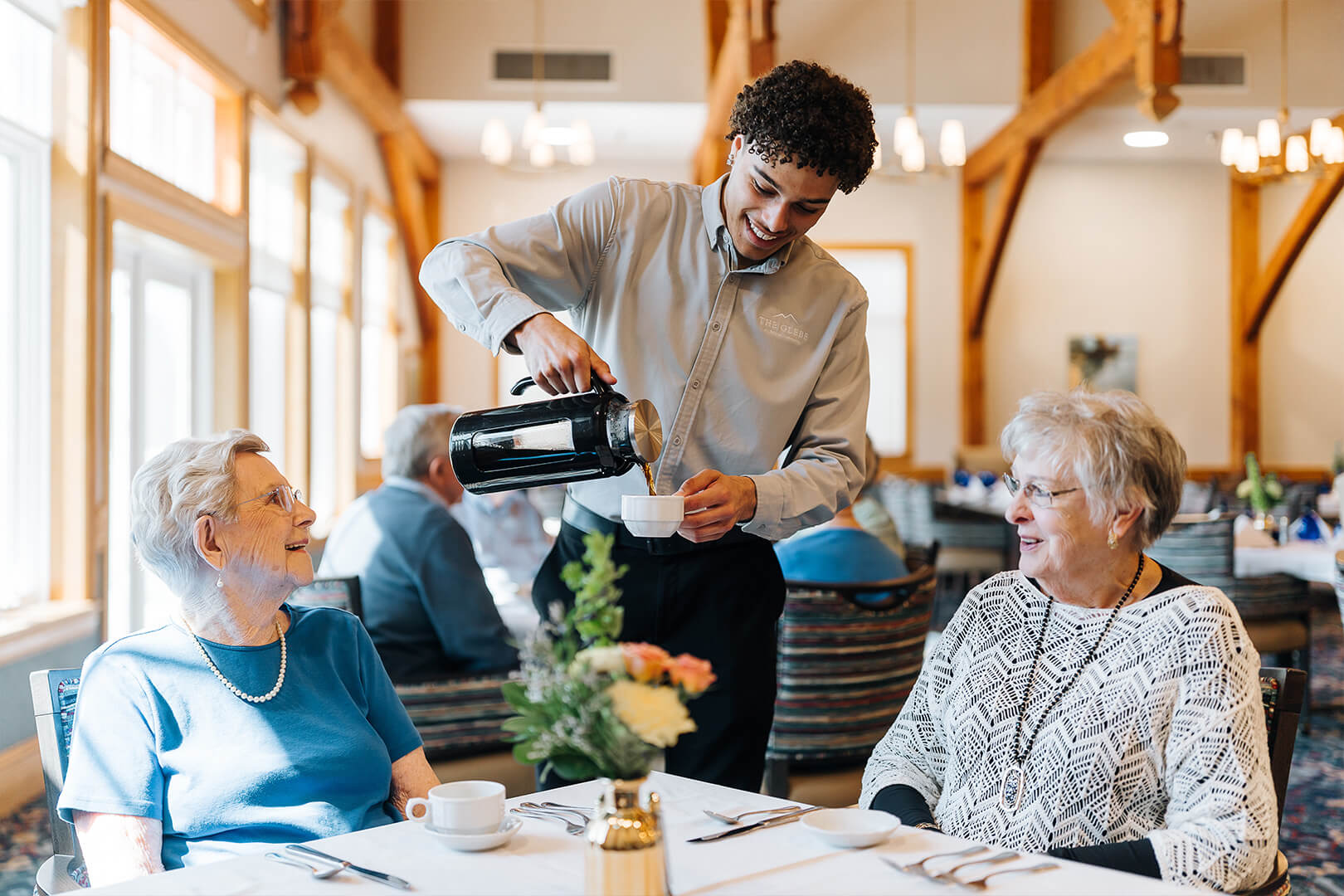 Smiling server pours coffee for two women in a bright dining area with wooden beams.