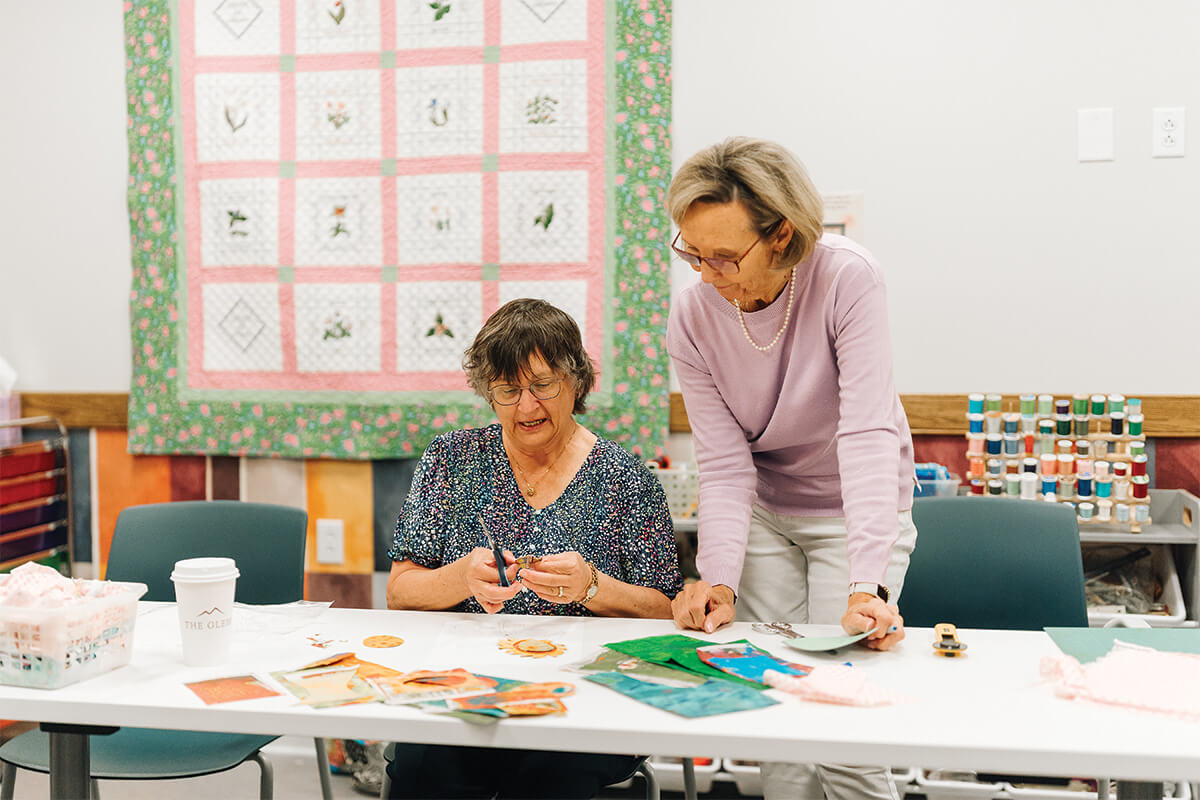 Two women working on quilting crafts at a table with materials and quilt on wall.