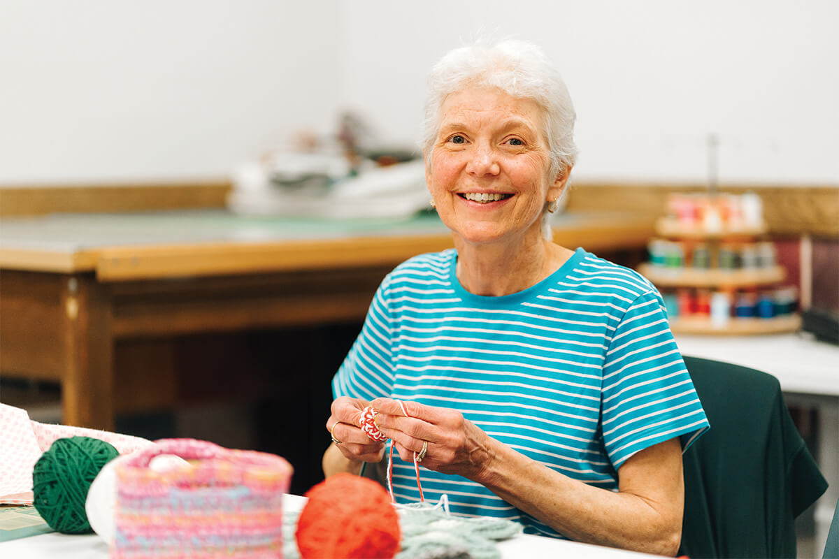 Smiling older woman knitting in a craft room with yarn on the table.