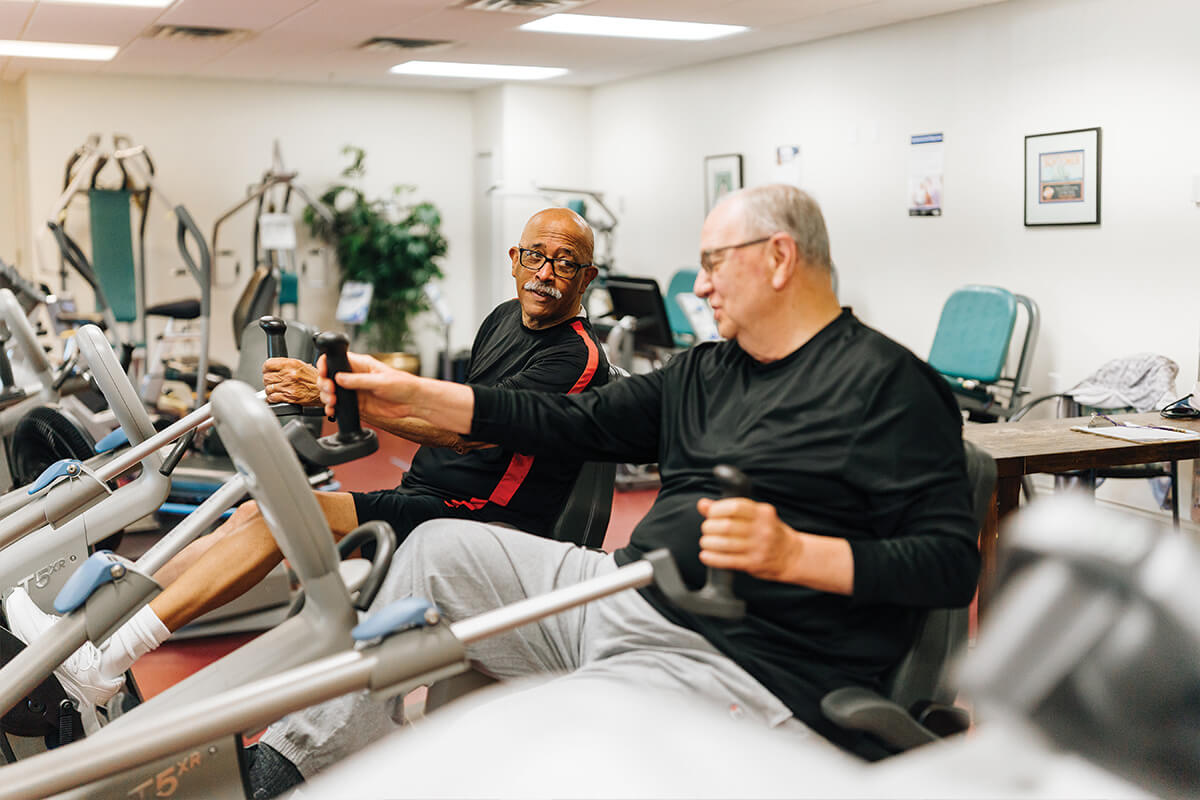 Two men exercising on machines in a fitness room at a senior living community.