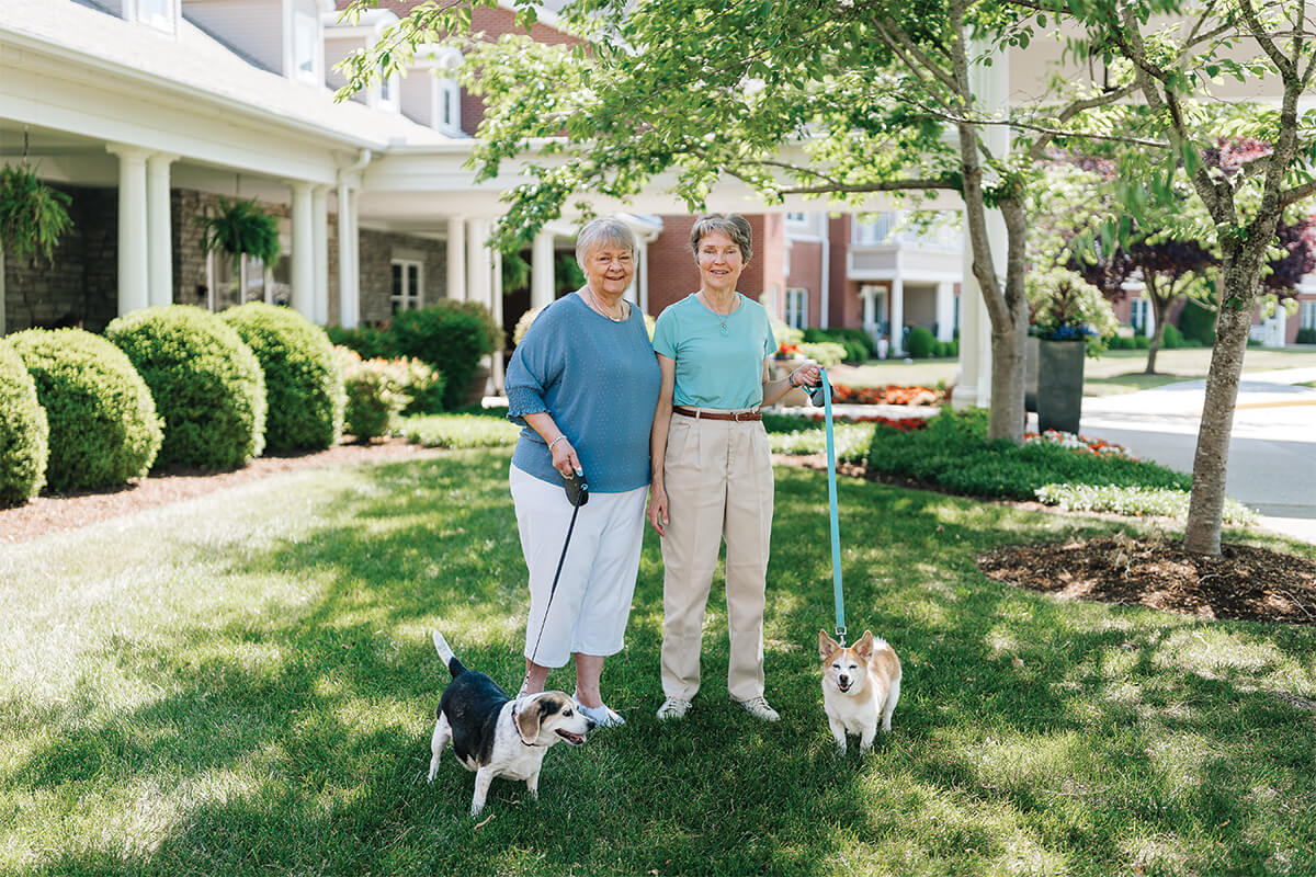 Two women walking dogs on a sunny day at a senior community courtyard.