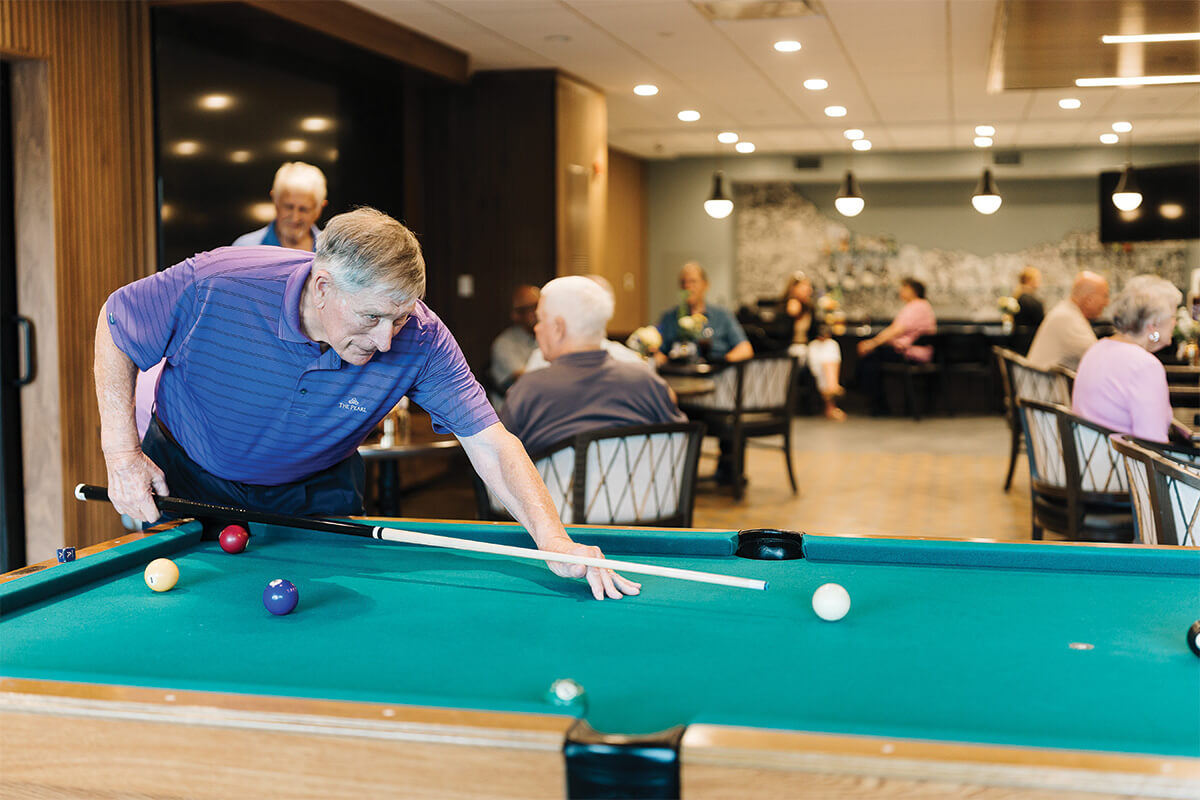 Resident playing pool in a vibrant common area with other senior community members.